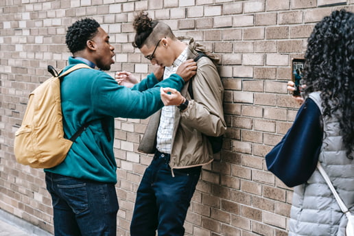 Two young men having a scuffle against a wall while a girl is recording with her phone