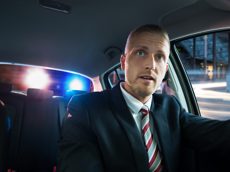 Man wearing a suit is being pulled over in his car with flashing blue and red lights behind him
